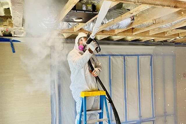 Technician in full PPE abrasive-blasting mold off attic rafters with a media hose inside tarped containment during a Green Restoration structural timber cleaning in the tri-state