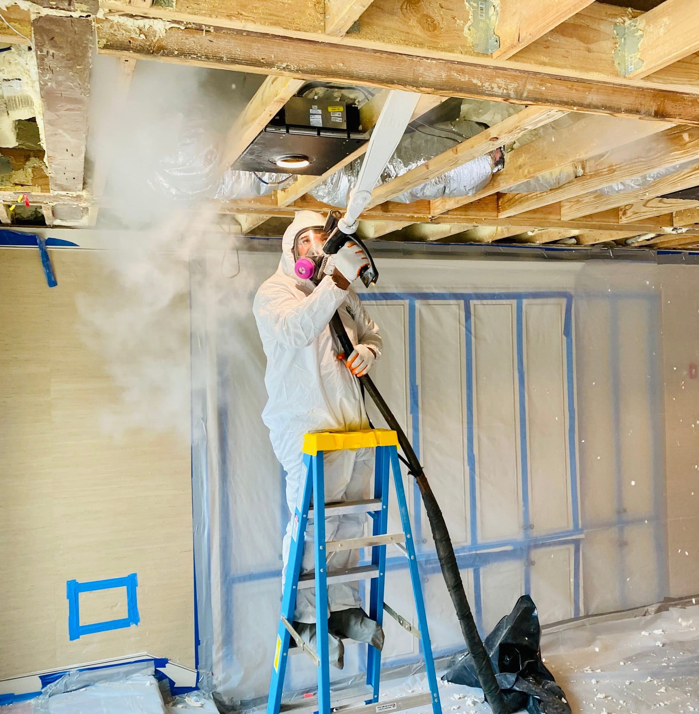 Green Restoration technician in full Tyvek PPE and respirator on a ladder treating a residential attic with visible mold growth across the roof sheathing and rafters during an active IICRC S520 remediation