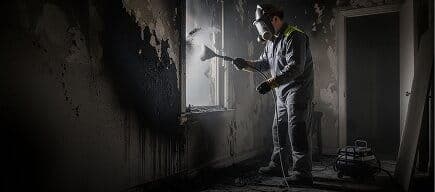 IICRC certified technician in protective coveralls wet sponging oily brown soot off a white painted living room wall, with a HEPA air scrubber and labeled content pack out bins visible in the background of a Connecticut residence