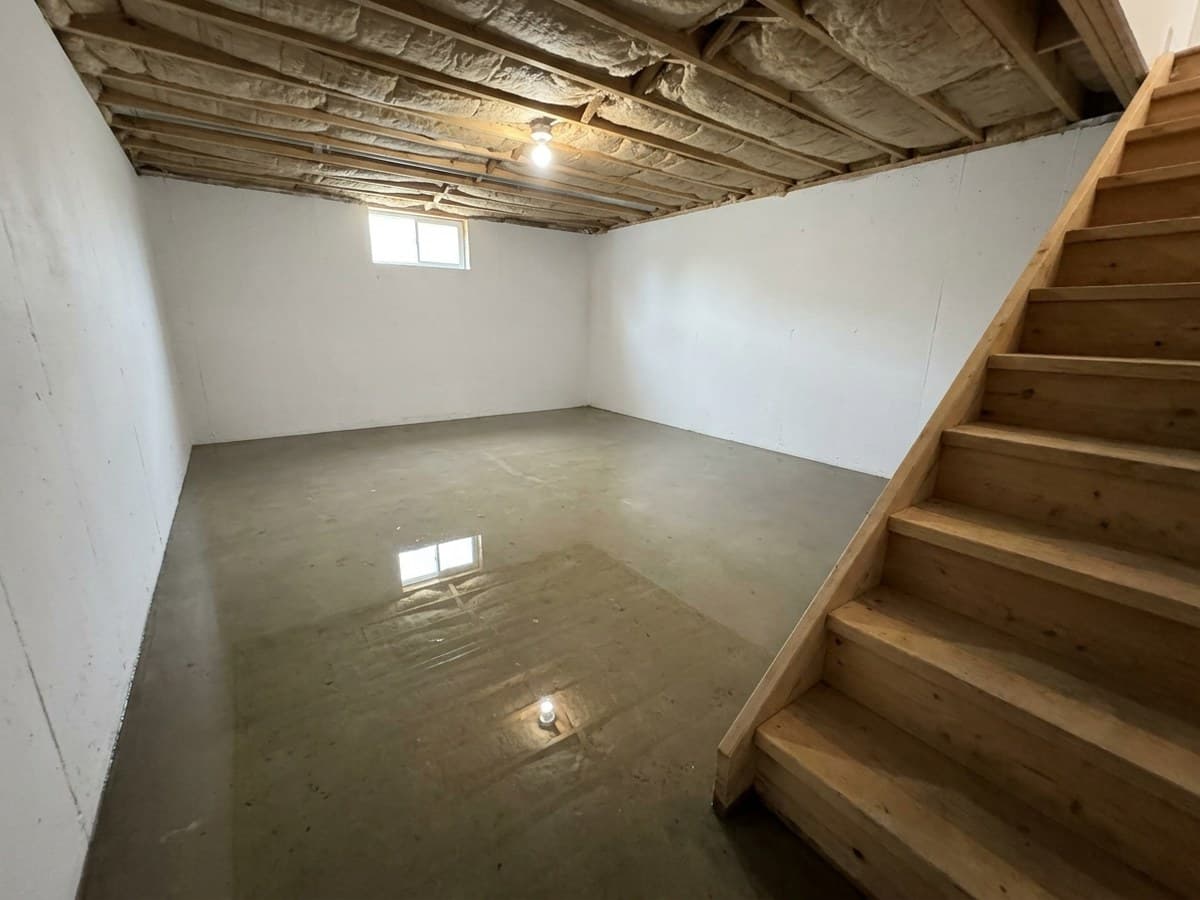Unfinished residential basement with a sheet of standing water across the concrete floor, a wooden staircase, and exposed floor joists overhead after a basement flood