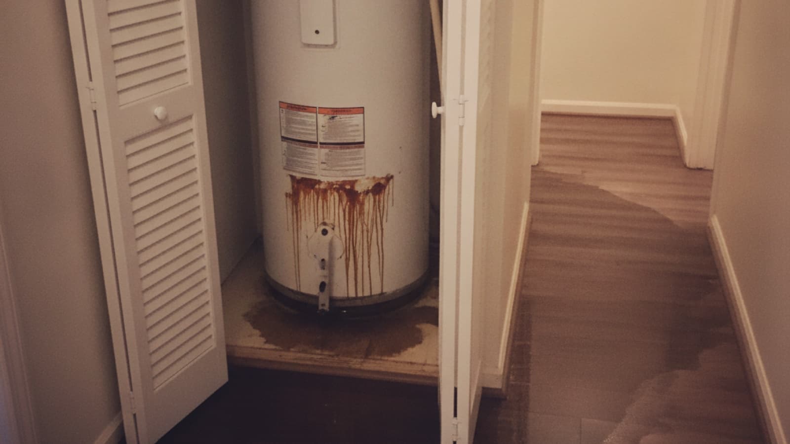 Water leaking from the base of a water heater in a hallway closet and spreading across the wood floor
